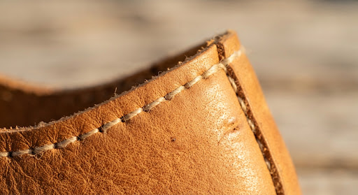 Extreme close up macro shot of handcrafted double-needle stitching along the curved edge of a minimal leather barefoot shoe, showing natural grain textures and organic plant-based thread in warm golden sunlight.