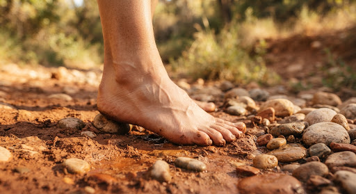 Close up photograph of a bare human foot grounded on sunlit red earth and smooth river rocks, capturing the tactile connection between natural human anatomy and organic terrain, warm warm tones of terracotta, clay, and sand, diffused golden afternoon light, soft focus natural background.
