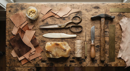 Overhead flatlay photograph of shoemaking hand tools on a weathered wooden workbench: antique brass shears, bone edge folder, organic beeswax block, natural vegetable leather offcuts, warm rustic artisan feeling.